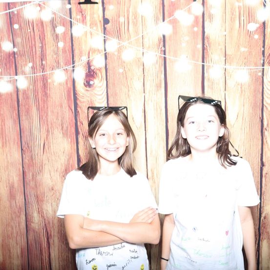 Zwei Mädchen stehen nebeneinander vor einer Holzwand mit Lichterketten. Sie tragen beide weiße T-Shirts.