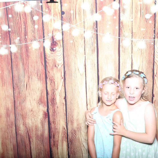 Zwei junge Mädchen stehen nebeneinander vor einer Holzwand mit einer Lichterkette. Das Mädchen auf der linken Seite hat eine Blumenkopfband.