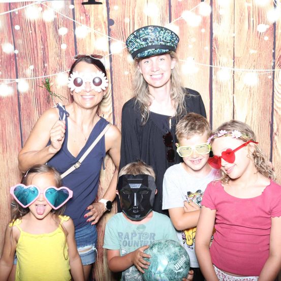 Eine Familie mit sechs Personen posiert für ein Foto vor einem hölzernen Hintergrund. Zwei Frauen tragen Sonnenbrillen und eine Mütze. Zwei Jungen tragen Masken und ein T-Shirt. Ein Mädchen trägt herzförmige Brillen.