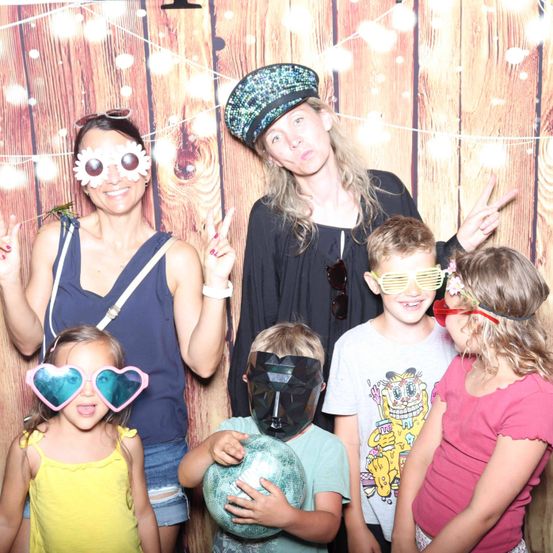 Eine Familie posiert für ein Foto vor einem hölzernen Hintergrund, alle tragen Sonnenbrillen. Eine Frau auf der linken Seite lächelt mit Blumen auf ihren Sonnenbrillen. Die Frau in der Mitte trägt eine Mütze und Sonnenbrille. Die Kinder tragen verschiedenfarbige Brillen.
