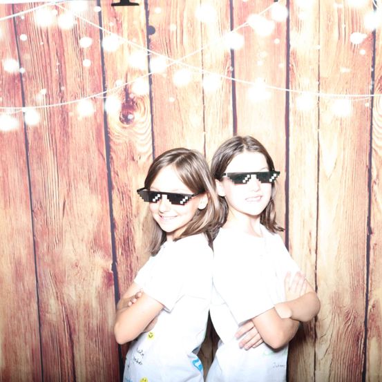 Zwei junge Mädchen in weißen T-Shirts und pixeligen Sonnenbrillen posieren vor einem hölzernen Hintergrund mit Lichterketten.