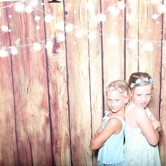 Zwei junge Mädchen mit Blumenkränzen stehen zusammen vor einem hölzernen Hintergrund mit Lichterketten.