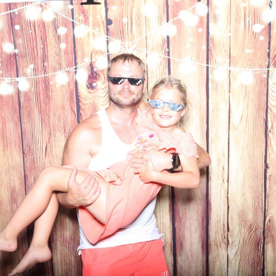 Ein Mann in Sonnenbrille hält ein lächelndes junges Mädchen in seinen Armen vor einem hölzernen Hintergrund.