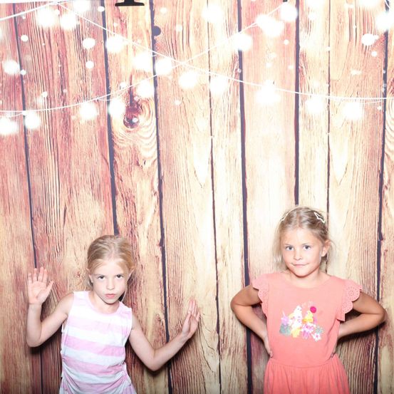 Zwei junge Mädchen posieren für ein Foto vor einem hölzernen Hintergrund mit dekorativen Lichterketten. Ein Mädchen trägt ein rosa Kleid mit Blumendesign.