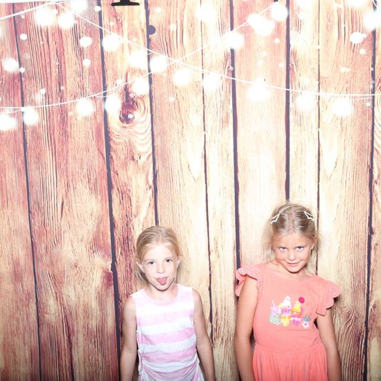 Zwei junge Mädchen stehen vor einem hölzernen Hintergrund mit dekorativen Lichtern. Das Mädchen auf der linken Seite hat ihre Zunge herausgestreckt. Das Mädchen auf der rechten Seite trägt ein rosa Kleid mit einem Cupcake-Design.