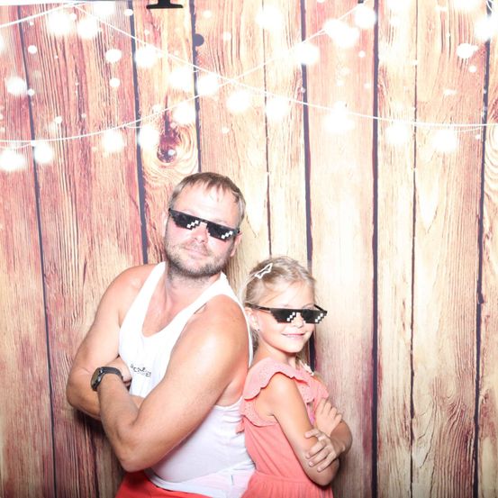 Ein Mann und ein junges Mädchen stehen lächelnd eng beieinander vor einem Holzhintergrund mit Lichterketten. Der Mann trägt ein weißes Tanktop und das Mädchen hat ein rosa Kleid an.