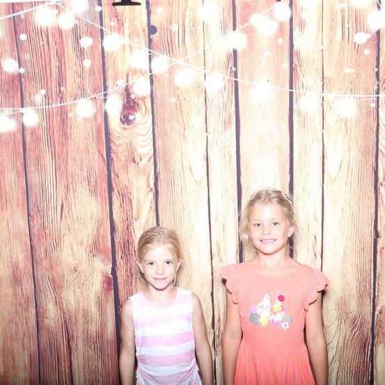 Zwei junge Mädchen stehen vor einer Holzwand mit Lichterketten. Sie lächeln für die Kamera.