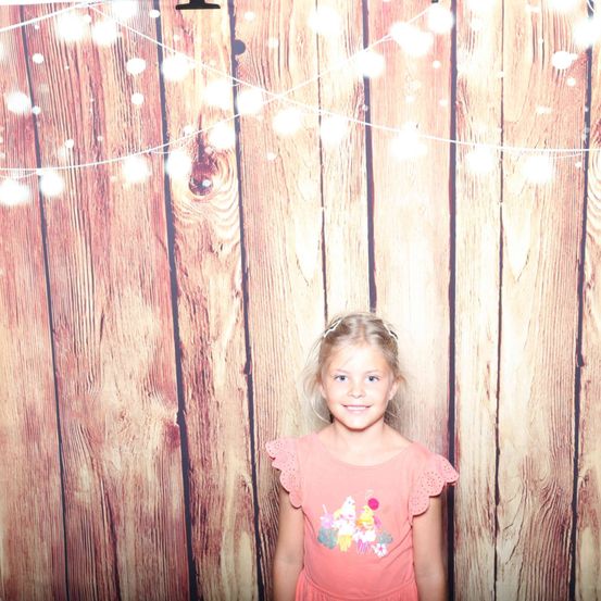 Ein kleines Mädchen steht vor einer hölzernen Kulisse mit dekorativen Lichtern darüber. Sie trägt ein rosa Kleid mit einem bunten Grafikdesign.
