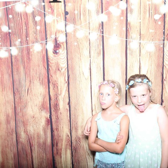 Zwei junge Mädchen stehen nebeneinander vor einer Holzwand mit Lichtern und Lichterketten. Sie tragen beide Sommerkleider und haben Blumen im Haar. Ein Mädchen streckt ihre Zunge heraus.