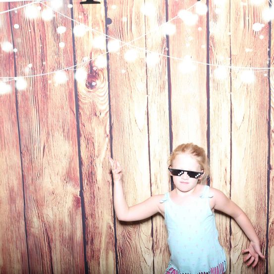 Ein junges Mädchen steht vor einem mit Lichtern geschmückten Holzhintergrund. Sie trägt ein hellblaues Tanktop und pixelierte Sonnenbrille. Ihre Hand ist erhoben, und sie schaut nach vorne.