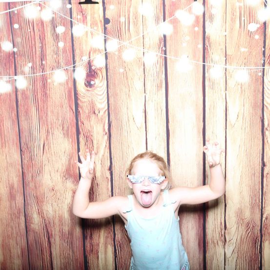 Ein kleines Mädchen steht vor einer Holzwand, trägt ein hellblaues Tanktop und weiße Brille. Sie hat den Mund offen und die Hände erhoben, als würde sie für ein Foto posieren.