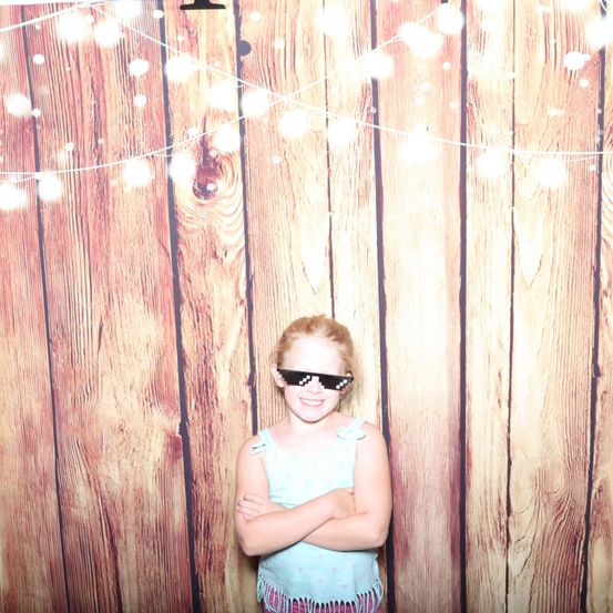 Ein junges Mädchen steht vor einer mit Lichterketten geschmückten Holzwand. Sie trägt ein hellblaues Tanktop und schwarze Sonnenbrille. Sie hat die Arme verschränkt.