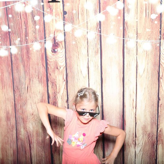 Ein kleines Mädchen in einem rosa Kleid und Pixelbrille posiert vor einem hölzernen Hintergrund mit weißen Lichtern.