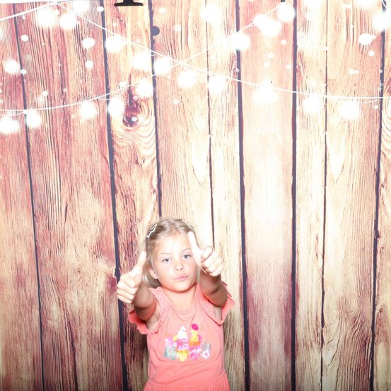 Ein kleines Mädchen in einem rosa Kleid zeigt den Daumen nach oben vor einem hölzernen Hintergrund mit Lichterketten.