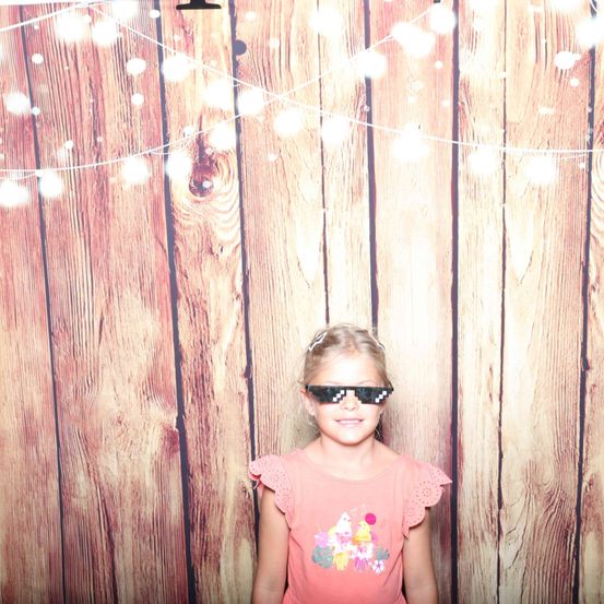 Ein kleines Mädchen steht vor einer mit Lichterketten geschmückten Holzwand und trägt ein rosa Kleid und pixelierte Sonnenbrille.
