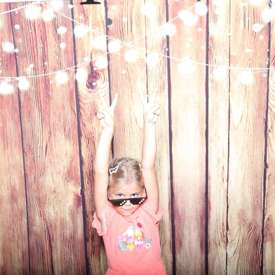 Ein kleines Mädchen in einem rosa Kleid und Sonnenbrille steht vor einem hölzernen Hintergrund mit Lichterketten. Sie hebt die Hände in einem Friedenszeichen.