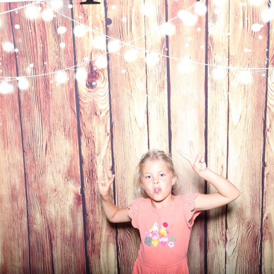 Ein kleines Mädchen mit blonden Haaren posiert vor einer mit Lichterketten dekorierten Holzwand. Sie macht ein Friedenszeichen mit beiden Händen.