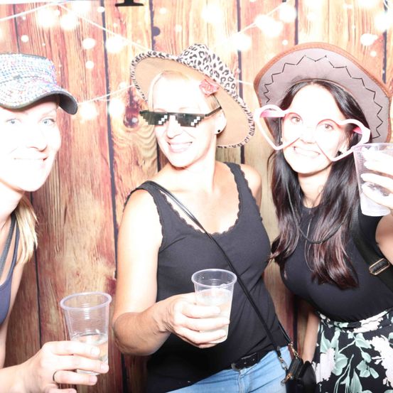 Drei Frauen in Hüten posieren mit Gläsern in den Händen für ein Foto vor einem hölzernen Hintergrund mit Lichtern.