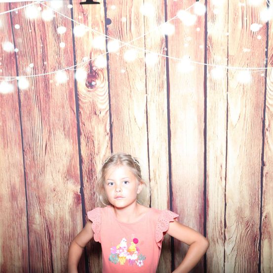 Ein junges Mädchen steht vor einem hölzernen Hintergrund mit Lichterketten. Sie trägt ein rosa Shirt mit einem bunten Cupcake-Design. Sie hat die Hände auf die Hüften gestellt und schaut direkt in die Kamera.