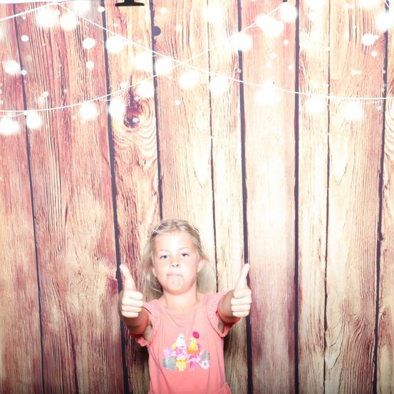 Ein junges Mädchen mit blonden Haaren steht vor einer Holzwand und gibt mit beiden Händen einen Daumen hoch. Die Wand hat Lichter und eine Lichterkette.