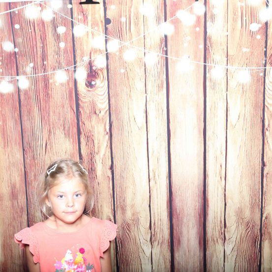 Ein kleines Mädchen steht vor einem hölzernen Hintergrund mit Lichterketten. Sie trägt ein rosa Shirt mit einem Eisdesign.