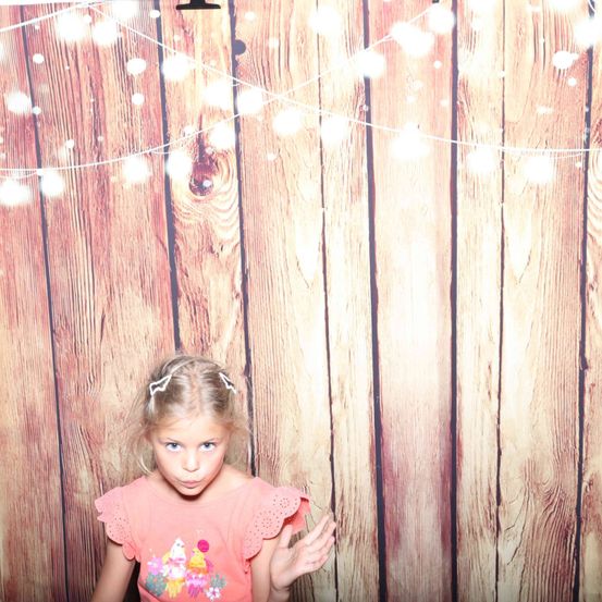 Ein kleines Mädchen mit blonden Haaren steht vor einer Holzwand mit dekorativen Lichtern und trägt ein rosa Oberteil mit einem bunten Cupcake-Design.