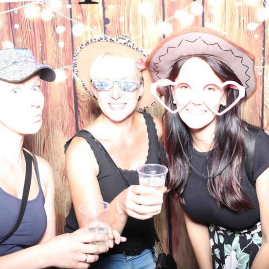 Drei Frauen posieren mit Getränken in der Hand und festlichen Hüten vor einem hölzernen Hintergrund für ein Foto.