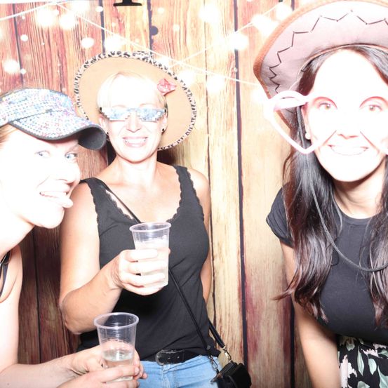 Drei Frauen lächeln für ein Foto in einem Fotokabinen. Die Frau in der Mitte hält ein Getränk. Alle tragen Hüte und einige haben Brillen. Ein hölzerner Hintergrund ist hinter ihnen.