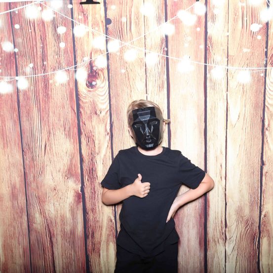 Eine Person in einem schwarzen Shirt und Maske steht vor einer Holzwand mit dekorativen Lichtern.