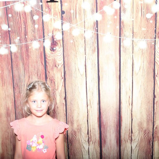Ein kleines Mädchen mit blonden Haaren steht vor einer Holzwand mit hängenden Lichterketten. Sie trägt ein rosa Shirt mit Cupcake-Design.