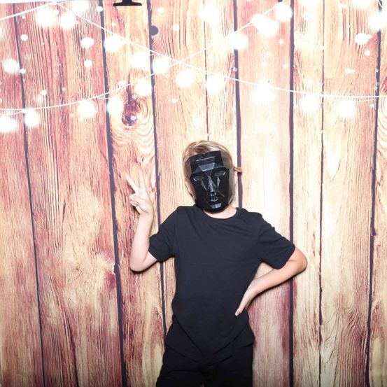 Eine Person in einem schwarzen T-Shirt und Maske steht vor einer Holzwand mit Lichterketten.
