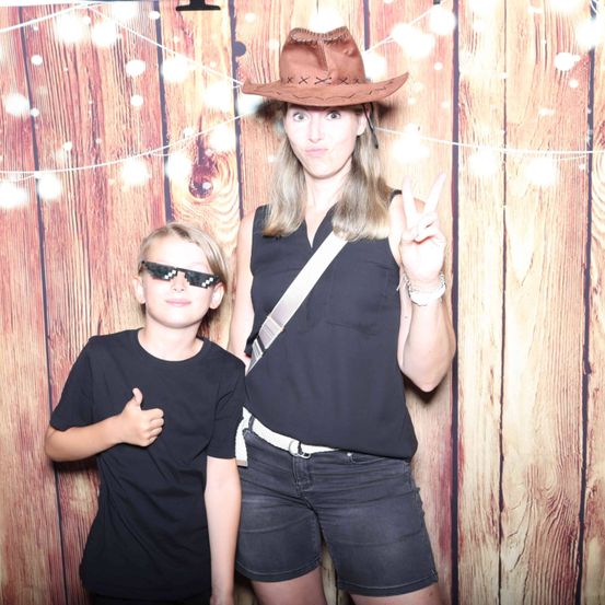Eine Frau mit Cowboyhut und Sonnenbrille macht ein Friedenszeichen, während ein Junge in einem schwarzen T-Shirt Daumen hoch in der Hand hat, vor einer Holzwand.