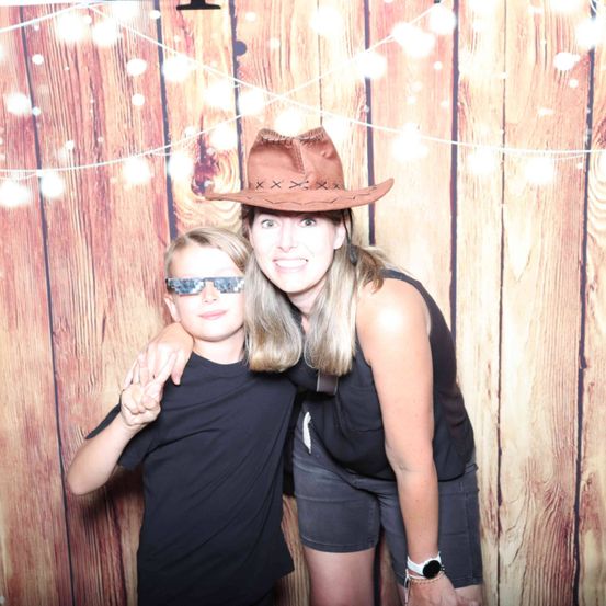 Eine Frau und ein junger Junge posieren für ein Foto vor einem hölzernen Hintergrund. Die Frau trägt einen Cowboyhut und der Junge eine Sonnenbrille.