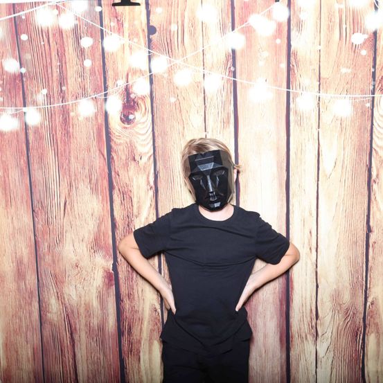 Eine Person mit schwarzer Maske steht vor einer Holzwand mit Lichterketten. Die Person trägt ein schwarzes T-Shirt und schwarze Hose.