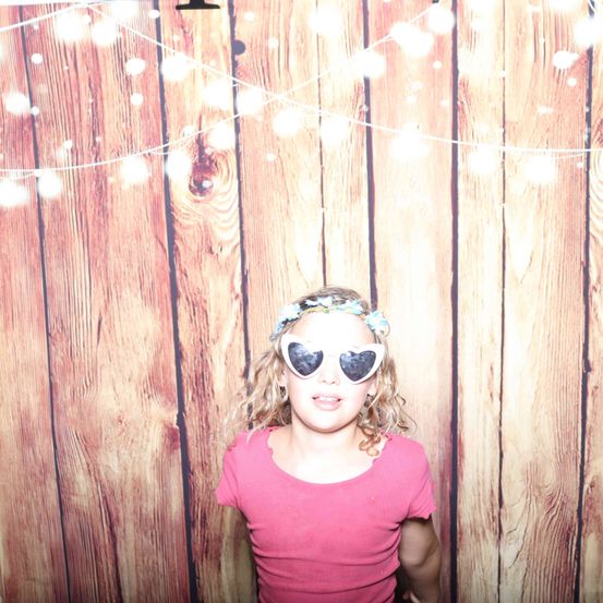 Ein kleines Mädchen mit herzförmigen Brillen und einem rosa T-Shirt steht vor einem hölzernen Hintergrund mit Lichterketten.