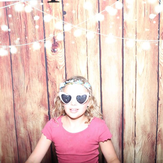 Ein kleines Mädchen mit einem rosa Shirt und Herz-förmigen Sonnenbrillen steht vor einer Holzwand mit Lichterketten.