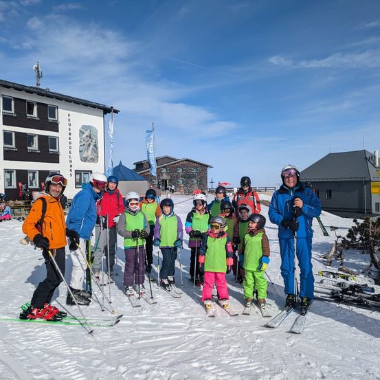 Eine Gruppe von Skifahrern, einschließlich Erwachsener und Kinder, posiert für ein Foto vor einem Gebäude mit einer Flagge an einem sonnigen Tag.