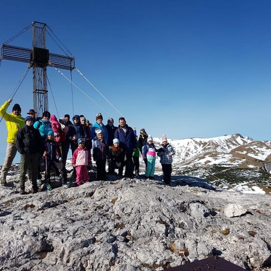 Eine Gruppe von Menschen steht auf einem Berggipfel und posiert für ein Foto vor einem großen Kreuz unter einem klaren blauen Himmel.