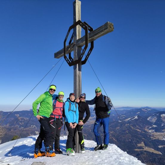 Vier Personen stehen auf einem verschneiten Gipfel und posieren für ein Foto vor einem großen Holzkreuz mit einer Metallstruktur oben. Das Wetter ist klar und die umliegenden Berge sind sichtbar.