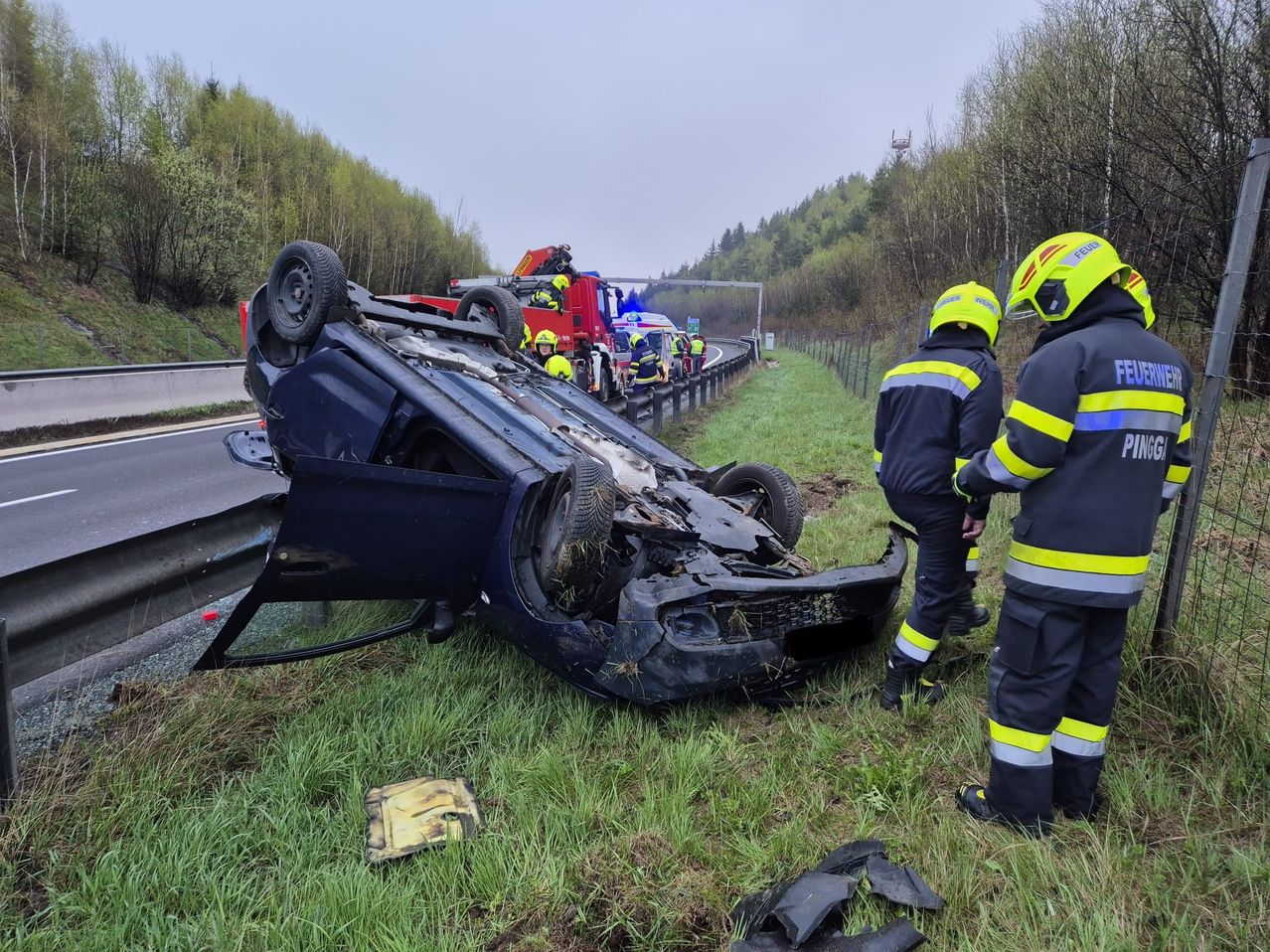 Ein demoliertes Auto liegt auf der Seite im Gras neben der Straße. Mehrere Feuerwehrleute tragen gelbe Helme.