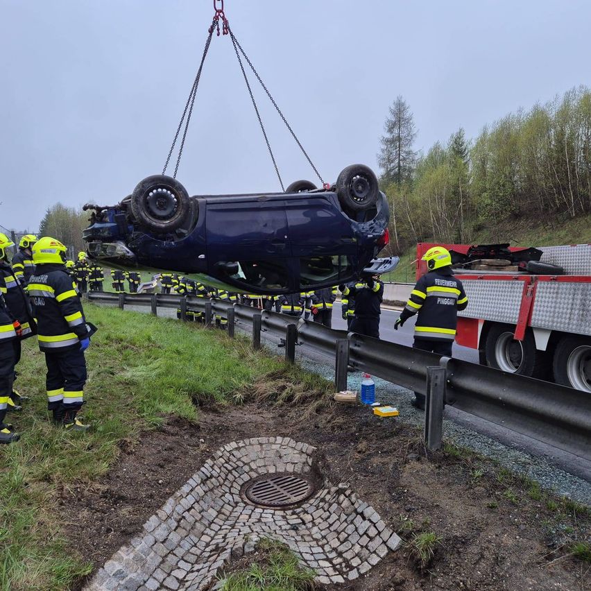 Ein blaues Fahrzeug wird von Feuerwehrleuten an einem Haken hochgezogen, während andere am Straßenrand stehen.