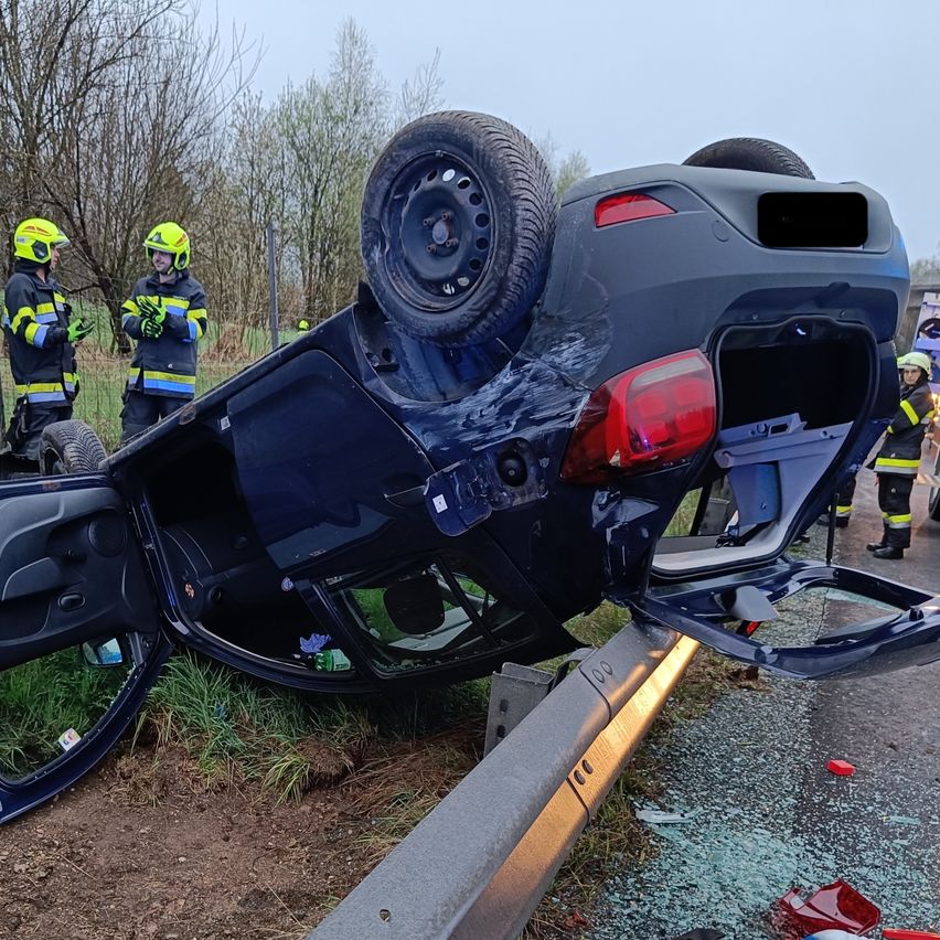 Ein dunkelblaues Auto hat sich auf die Seite gedreht und ruht am Straßenrand. Das Hinterreifen ist noch befestigt. Zwei Feuerwehrleute in gelben Helmen inspizieren das Fahrzeug.