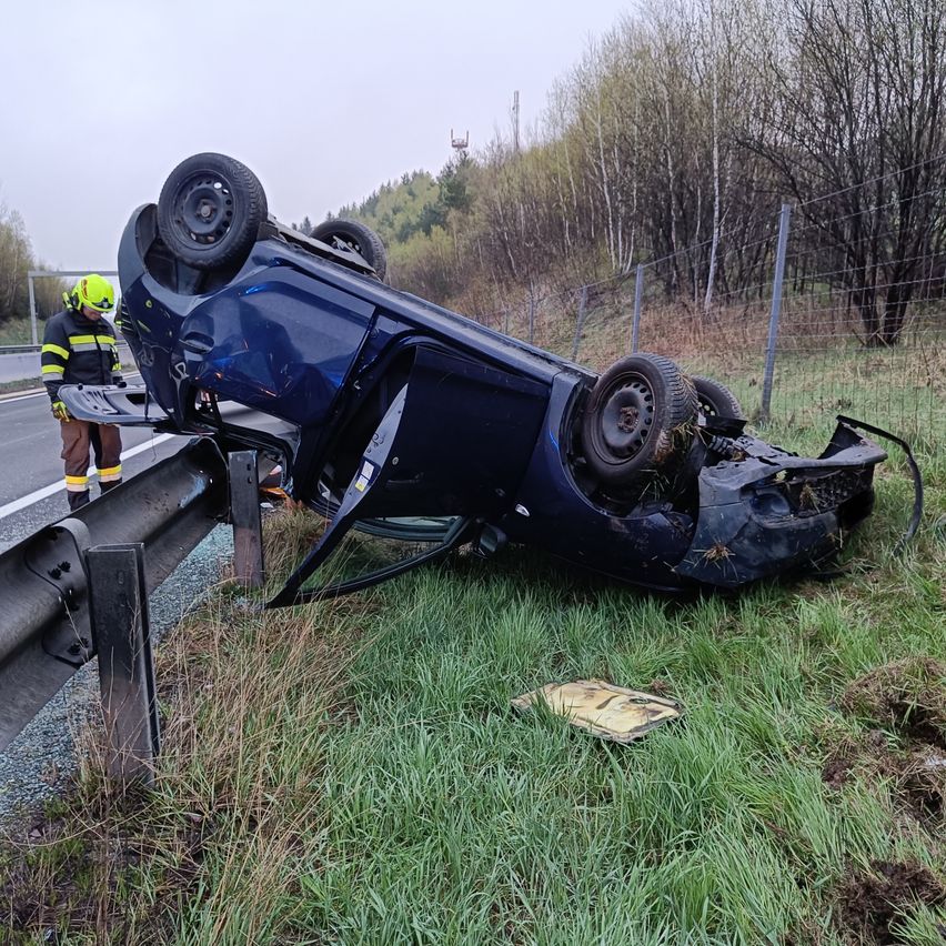 Ein blaues Auto ist auf der Straßenseite umgekippt, mit einem Rettungsarbeiter in der Nähe, der einen Helm und eine reflektierende Weste trägt.
