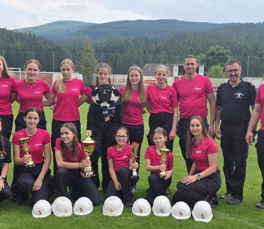 Eine Gruppe junger Frauen und zwei Männer in passenden rosa T-Shirts stehen auf einem Feld mit Bergen im Hintergrund. Sie halten Pokale und weiße Helme.