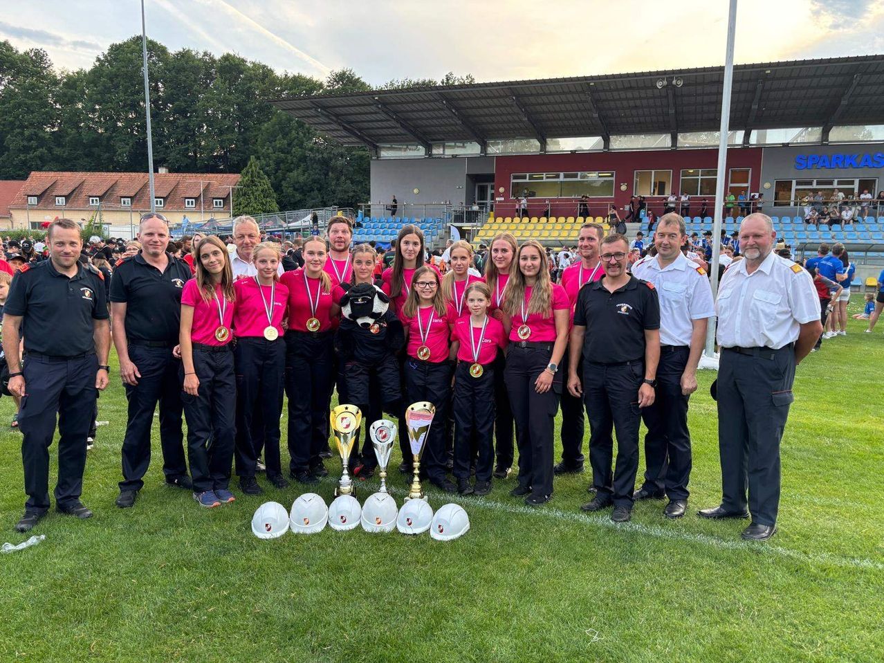 Ein Sportteam mit mehreren Frauen und Männern steht auf einem Feld, hält Medaillen und Trophäen und posiert für ein Foto. Sie tragen alle passende Uniformen.