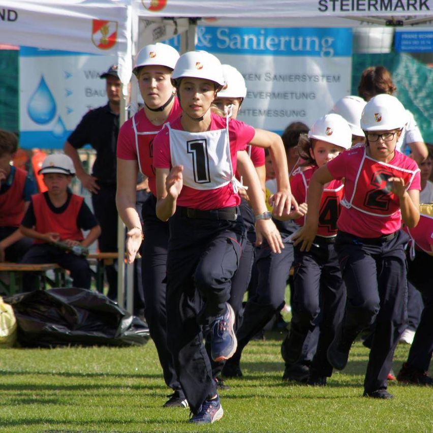 Eine Gruppe von Kindern in roten Uniformen und weißen Helmen läuft auf einem Rasenfeld, möglicherweise bei einem Rennen. Dahinter stehen Menschen in Hüten und Uniformen, von denen einer steht.