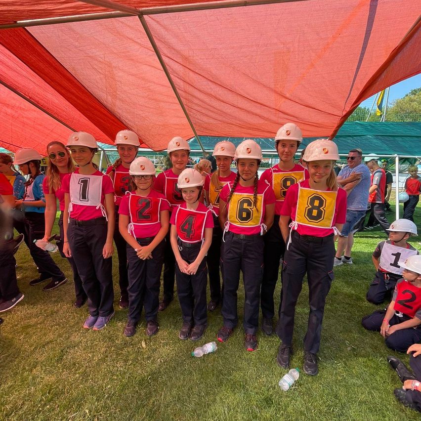 Eine Gruppe junger Mädchen in passenden Uniformen und nummerierten Trikots posiert für ein Foto unter einem roten Zelt. Sie tragen weiße Helme und einige haben eine Brille. Die Umgebung scheint ein Outdoor-Event zu sein.