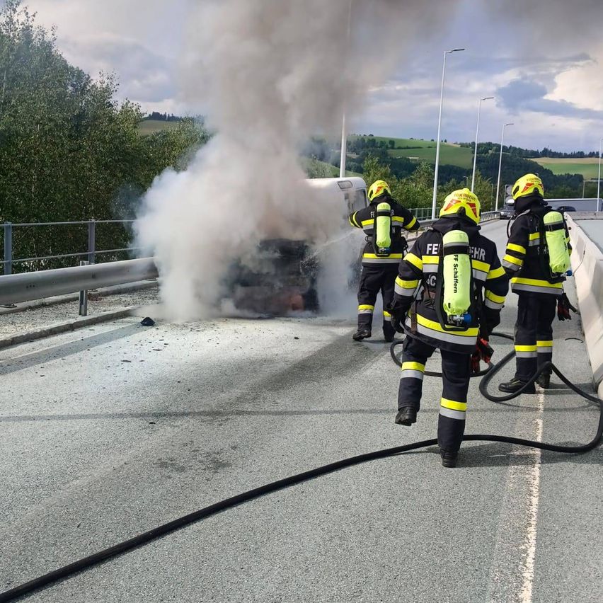 Feuerwehrleute löschen ein brennendes Fahrzeug am Straßenrand, wobei Rauch in die Luft aufsteigt.