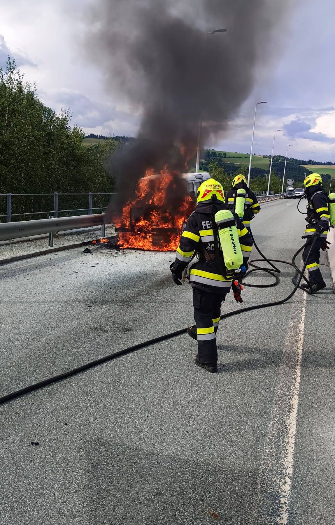 Feuerwehrleute in voller Ausrüstung löschen ein brennendes Fahrzeug auf einer Autobahn. Einer hält einen Schlauch, während andere helfen.