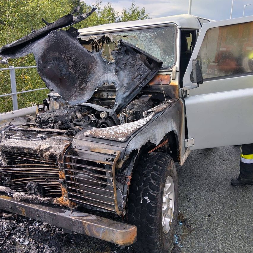 Ein ausgebranntes Fahrzeug mit beschädigter Motorhaube und offener Tür steht auf einer Straße mit einem Metallzaun. Bäume und ein bewölkter Himmel im Hintergrund.